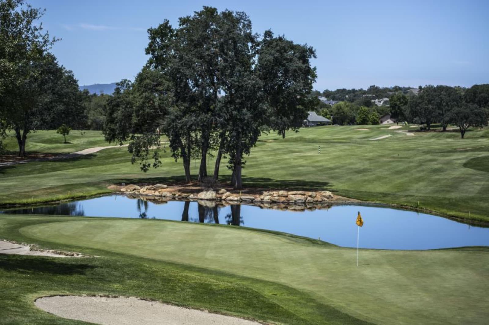 A golf green with a yellow pin flag beside a pond and oak trees