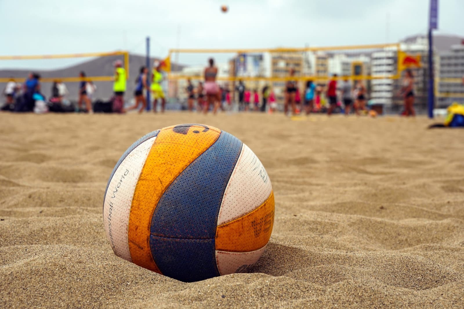 A volleyball resting in the sand with a beach game in the background